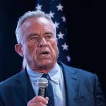 Man holding microphone, speaking with American flag behind.