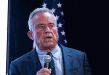 Man holding microphone, speaking with American flag behind.