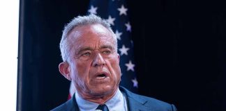 Man holding microphone, speaking with American flag behind.