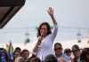 Woman speaking at outdoor event with raised hand.