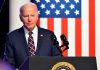 Man in suit speaking at podium with American flag backdrop.