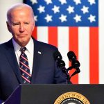 Man in suit speaking at podium with American flag backdrop.