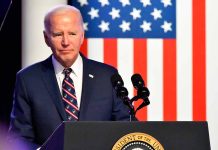 Man in suit speaking at podium with American flag backdrop.