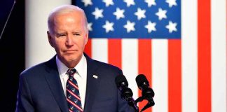 Man in suit speaking at podium with American flag backdrop.