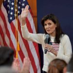 Woman speaking into microphone beside American flags.