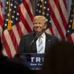 Person speaking at podium with American flags behind.