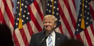 Person speaking at podium with American flags behind.