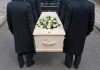 Four people carrying a coffin with white flowers.