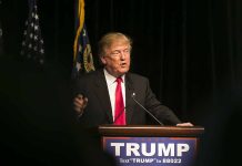 Man speaking at podium with "TRUMP" sign.