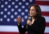 Woman speaking into microphone with American flag backdrop.