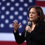 Woman speaking into microphone with American flag backdrop.