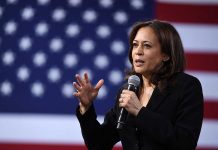 Woman speaking into microphone with American flag backdrop.