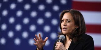 Woman speaking into microphone with American flag backdrop.