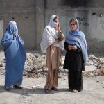 Three women in traditional attire standing in an urban setting with debris in the background