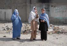 Three women in traditional attire standing in an urban setting with debris in the background