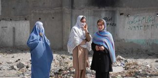 Three women in traditional attire standing in an urban setting with debris in the background