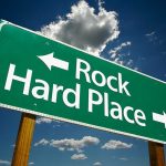 Directional road sign indicating Rock to the left and Hard Place to the right against a blue sky