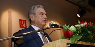 A man in a suit speaking at a podium with flowers and a microphone