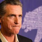 Newsom’s SHOCKING Biden Confession Rocks Democrats Man in suit solemn expression purple background with bear drawing