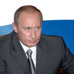 A serious looking man in a suit sitting at a table with a Russian flag in the background