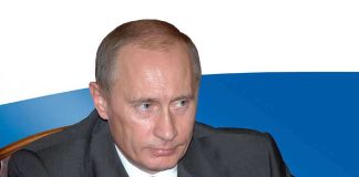 A serious looking man in a suit sitting at a table with a Russian flag in the background