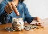 Person placing a coin into a savings jar labeled 'SAVINGS'