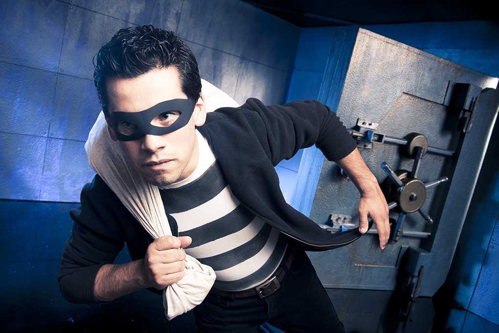 A masked thief in a striped shirt holding a bag while emerging from a bank vault