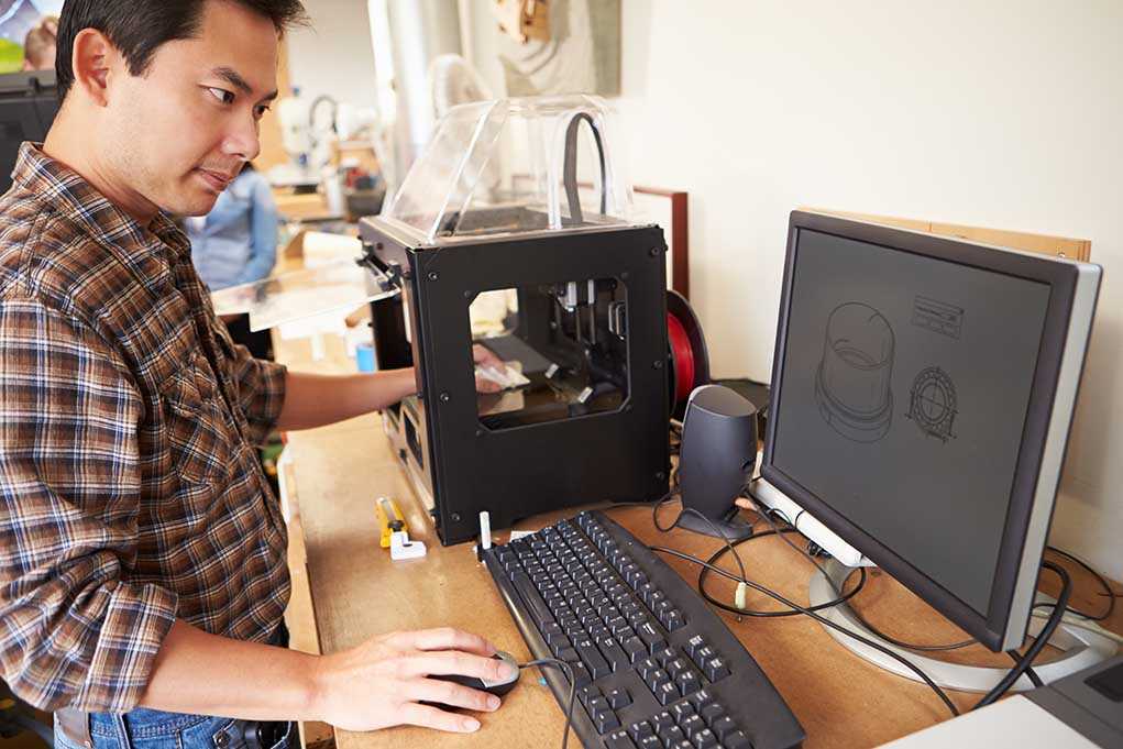 A person working at a desk with a 3D printer and computer