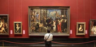 A person standing in an art gallery, observing a large historical painting surrounded by other artworks