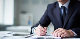 A man in a suit writing notes at a desk with documents and a tablet