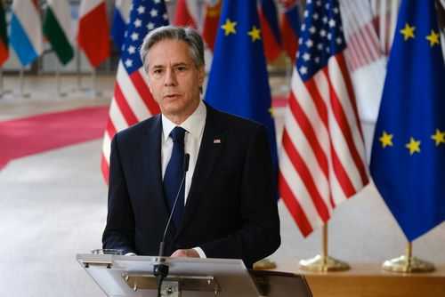 shutterstock_2284726273 Man in suit speaking at podium with international flags behind