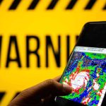 Tropical Tag, Hurricane-Size Havoc Hand holds smartphone showing cyclone radar map against warning sign