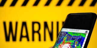 Tropical Tag, Hurricane-Size Havoc Hand holds smartphone showing cyclone radar map against warning sign