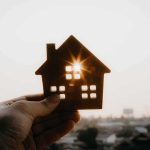 America’s Apartment Market Is Facing A Painful Reset Hand holding a small house cutout against a bright sunset sky