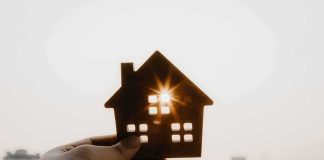 Hand holding a small house cutout against a bright sunset sky