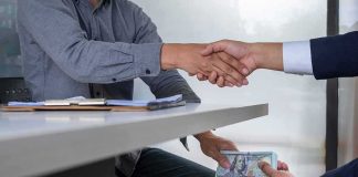 Two people shake hands over a desk while cash is exchanged underneath