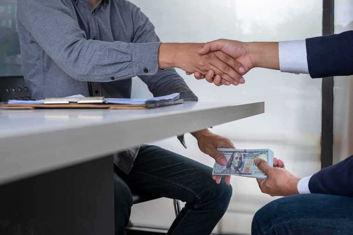 shutterstock_1169658241 Two people shake hands over a desk while cash is exchanged underneath