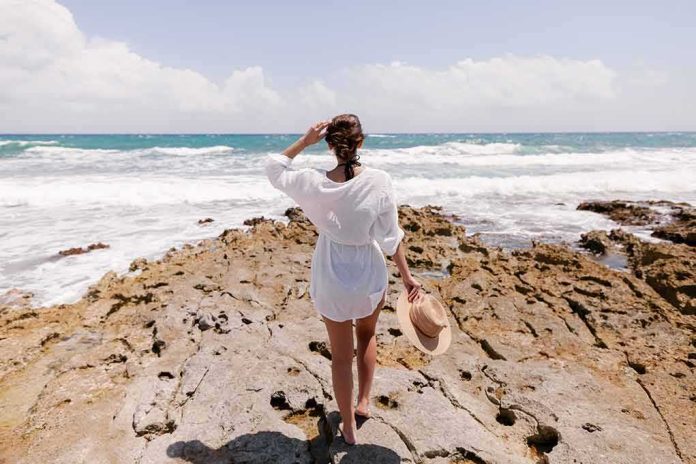 shutterstock_1682508124 Woman in white cover-up standing on rocky shore by the ocean