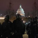 Military On Main Street — Legal Line Crossed? Riot police in protective gear stand before a domed capitol building at night