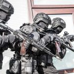Tactical officers in black gear stack up with rifles by a building