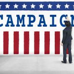 Propaganda Ban Tested Weeks Before Midterms Man in suit writing on large campaign banner with stars and stripes